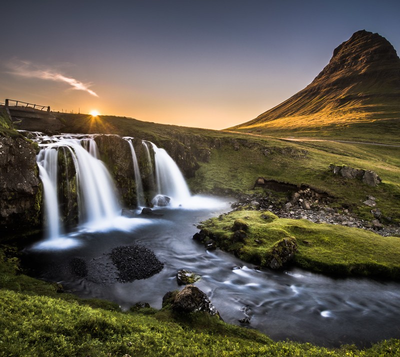 A waterfall in the middle of a grassy field with a mountain in the background (bridge, landscape, river, sunset)