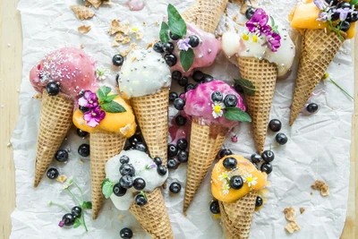 Conos de helado coloridos cubiertos con frutas frescas, flores comestibles y salsas, dispuestos artísticamente sobre papel pergamino arrugado.