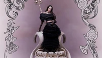 Jang Wonyoung exala elegância e poder em um dramático conjunto preto, sentada em um trono ornamentado cercado por intrincados designs florais.