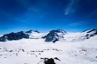 Cordilheira coberta de neve emergindo de um manto de nuvens sob um céu azul claro.