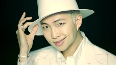 RM do BTS em um traje branco estiloso e chapéu, exibindo um sorriso carismático.