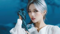 Soyeon do (G)I-DLE exibindo um visual marcante com cabelo trançado contra um fundo azul dramático.