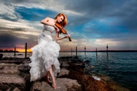 Una elegante violinista en un vestido blanco fluido toca su instrumento al atardecer junto a la orilla del agua, capturando la esencia de la belleza y la creatividad contra un cielo impresionante.