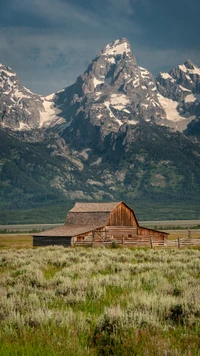 Grange rustique nichée dans une vaste prairie avec des montagnes majestueuses et des nuages en arrière-plan.