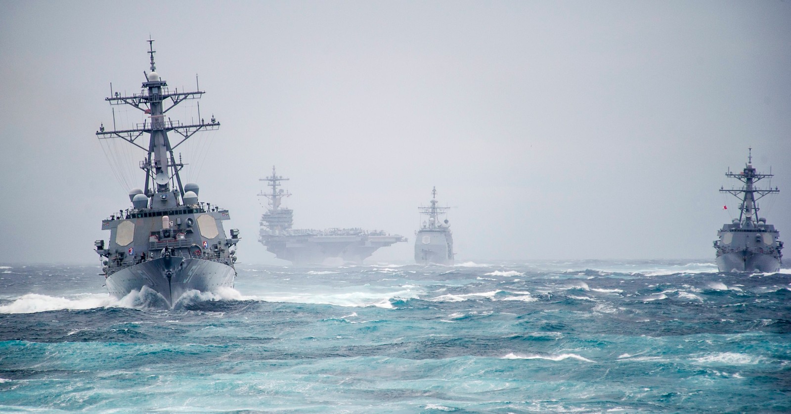 Vista aérea de barcos de guerra en el océano con algunos barcos más pequeños al fondo (grupo de ataque de portaaviones, marina de los estados unidos, portaaviones, destructor, buque de guerra)