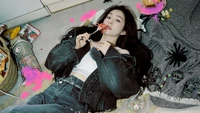 Irene from Red Velvet lies on a floor scattered with colorful snacks and toys, holding a lollipop, exuding a playful yet stylish vibe.