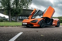 2021 McLaren 720S в ярком оранжевом цвете с крыльями-бабочками на фоне современного архитектурного пейзажа.