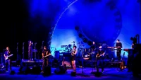 Dynamische Rockkonzertaufführung mit einem vielfältigen musikalischen Ensemble auf der Bühne, mit einem Schlagzeuger, Gitarristen und Sängern, vor einem lebhaften blauen Hintergrund.