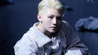 Woozi from Seventeen in a striking pose, showcasing a unique style with platinum blonde hair and a white outfit against a moody, icy background.