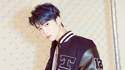 Jaehyun de NCT 127 dans une veste varsity élégante avec des cheveux bleus, posant devant une clôture en treillis métallique.