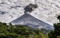 Erupting stratovolcano amidst a dramatic cloud-filled sky, surrounded by lush greenery.