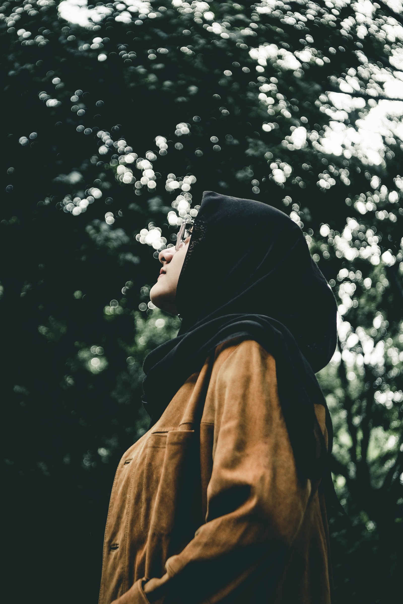 Mujer árabe con una chaqueta marrón y un hoodie negro de pie frente a los árboles (alá, allah, niña, hiyab, islam)