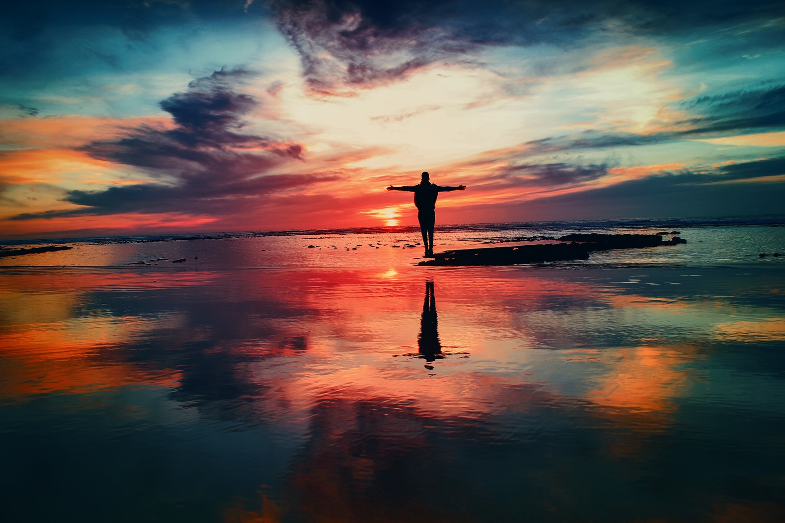 Um homem em pé na praia com os braços abertos no ar (reflexo, água, por do sol, horizonte, nuvem)