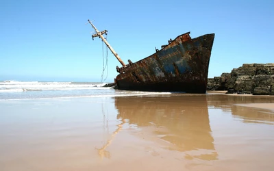 Épave abandonnée sur une rive sereine, se reflétant dans des eaux calmes sous un ciel bleu clair.