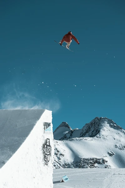 Um snowboarder realiza um truque aéreo acima de um penhasco nevado, contra um céu azul deslumbrante e um fundo de picos de montanha irregulares.