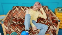 Yeonjun do TXT relaxando em um sofá de estilo retrô, exibindo um visual casual, mas estiloso, com cabelo em cores pastel e óculos da moda.