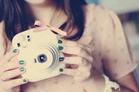 A girl holding a white camera adorned with a pink bow, showcasing her stylish manicure and soft beauty.