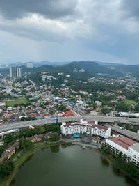 Vista aérea de uma área urbana residencial com um lago, vegetação exuberante e um fundo de montanhas sob um céu nublado.