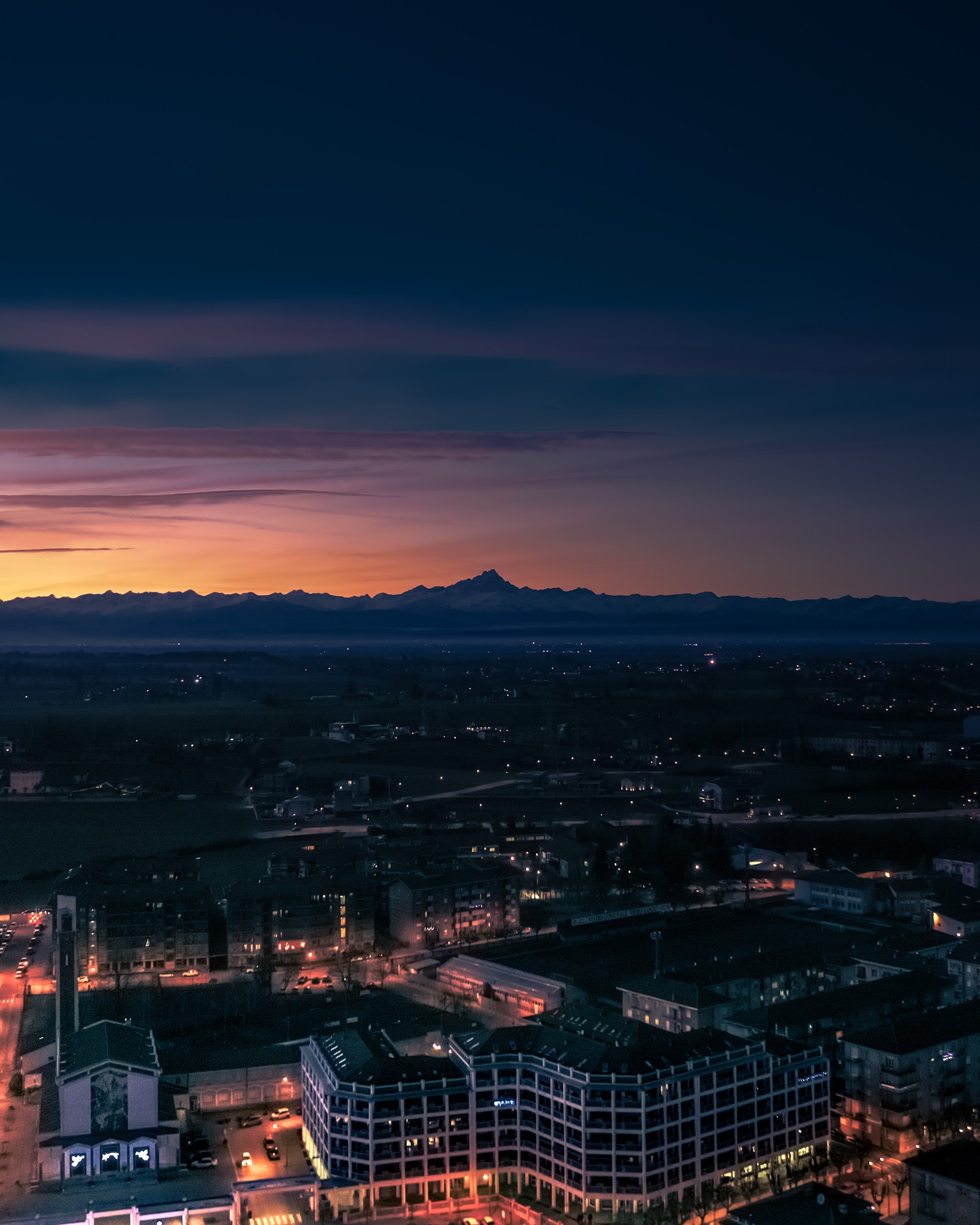 Une vue aérienne d'une ville la nuit avec une montagne au loin (atmosphère, nuage, bâtiment, crépuscule, immeuble tour)