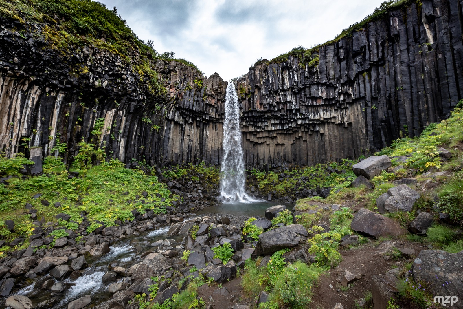 A waterfall flowing into a rocky gorge surrounded by green vegetation (waterfall, cloud, water, water resources, plant)