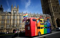 Um vibrante ônibus de dois andares adornado com bandeiras do arco-íris passa pelo icônico Palácio de Westminster, simbolizando diversidade e celebração no coração da cidade capital.