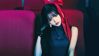 Jang Wonyoung exudes a captivating blend of elegance and attitude, seated against a backdrop of plush red seats, adorned with a striking red bow and stylish accessories.