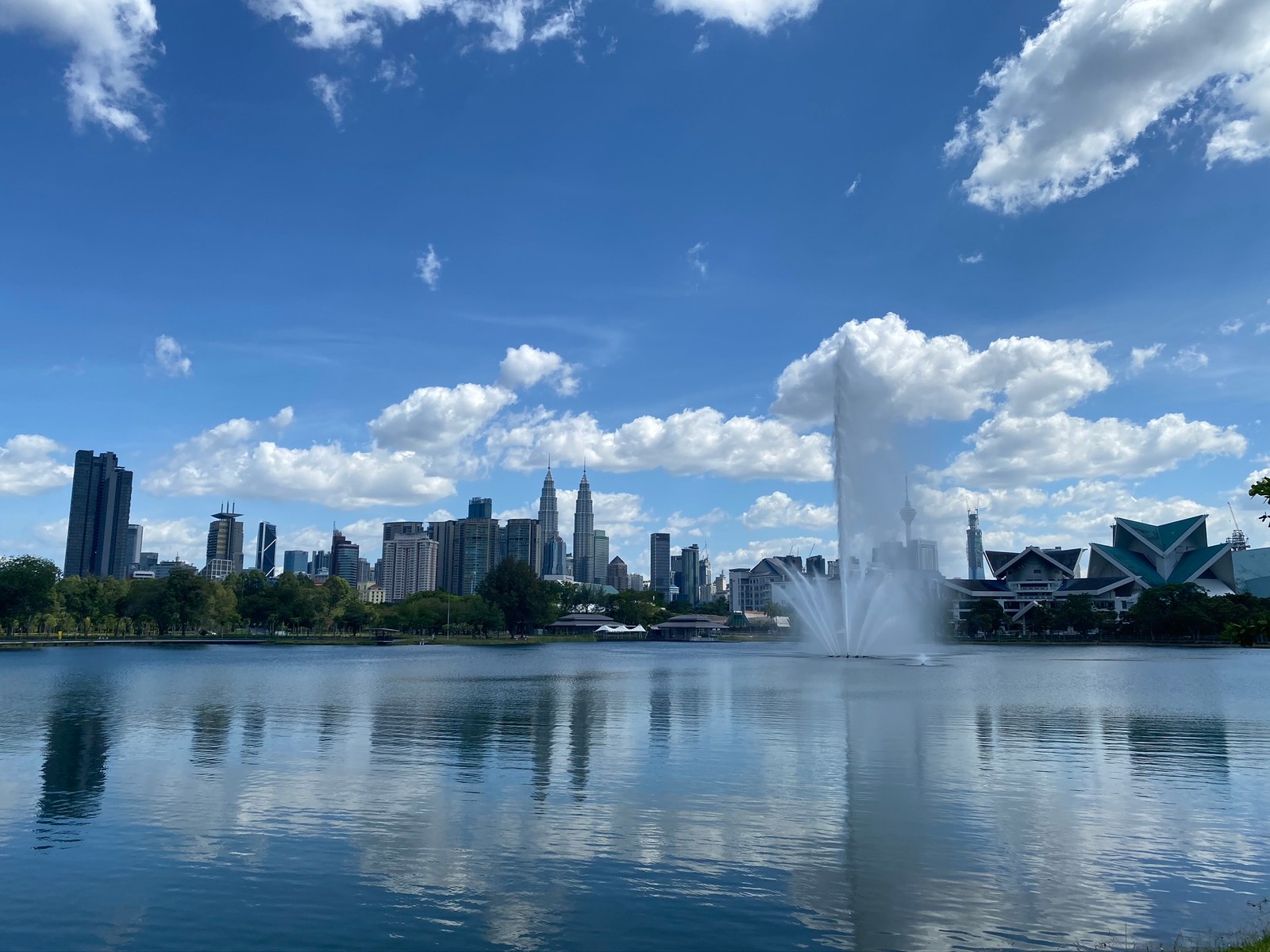 Uma linda fonte no meio de um lago com uma cidade ao fundo (torres petronas, kuala lumpur, recursos hídricos, água, corpo de água)