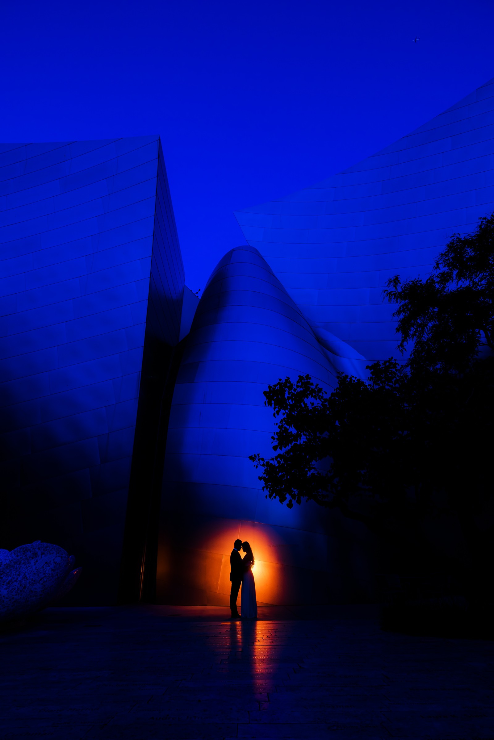 Il y a deux personnes debout devant un bâtiment la nuit (bleu, lumière, nuit, obscurité, arbre)