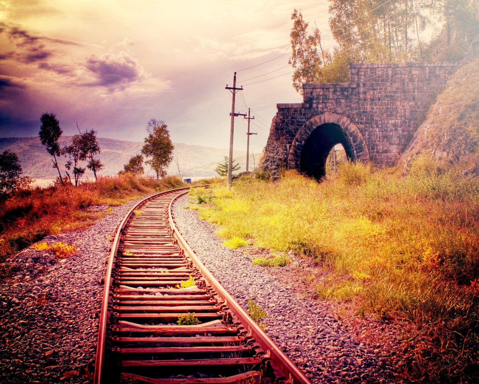 Há uma linha de trem passando por um túnel com uma ponte ao fundo (trilho, por do sol, trem, túnel)