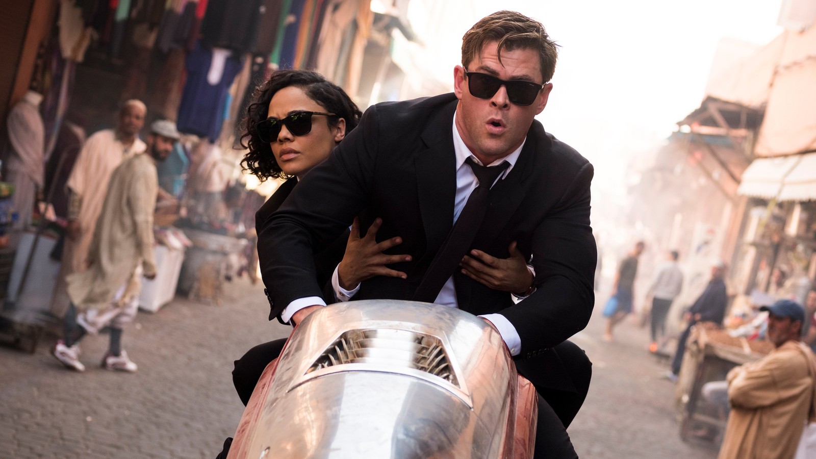 Imagen borrosa de un hombre en traje y gafas de sol montando una motocicleta con una mujer en traje (hombres de negro internacional, película, chris hemsworth, tessa thompson)