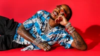 Stylish rapper lounging in vibrant blue graphic shirt against a bold red backdrop, showcasing unique fashion and eyewear.