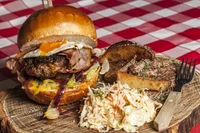 Hearty hamburger topped with bacon, egg, and melted cheese, served alongside crispy potato wedges and creamy coleslaw on a rustic wooden platter.