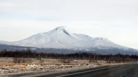 Snow-capped stratovolcano rising majestically over a winter landscape, framed by a winding road and sparse trees against a clear sky.