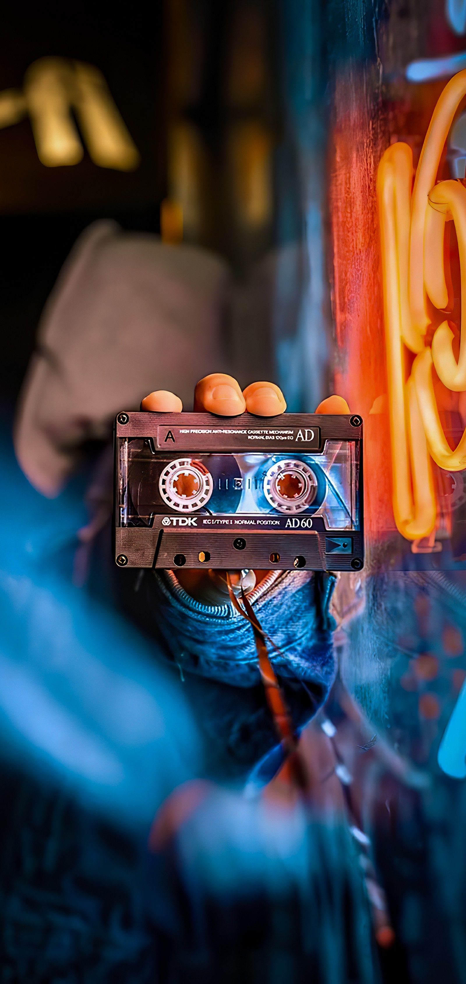 Someone holding a cassette in front of a neon sign (orange, purple, finger, gas, electric blue)