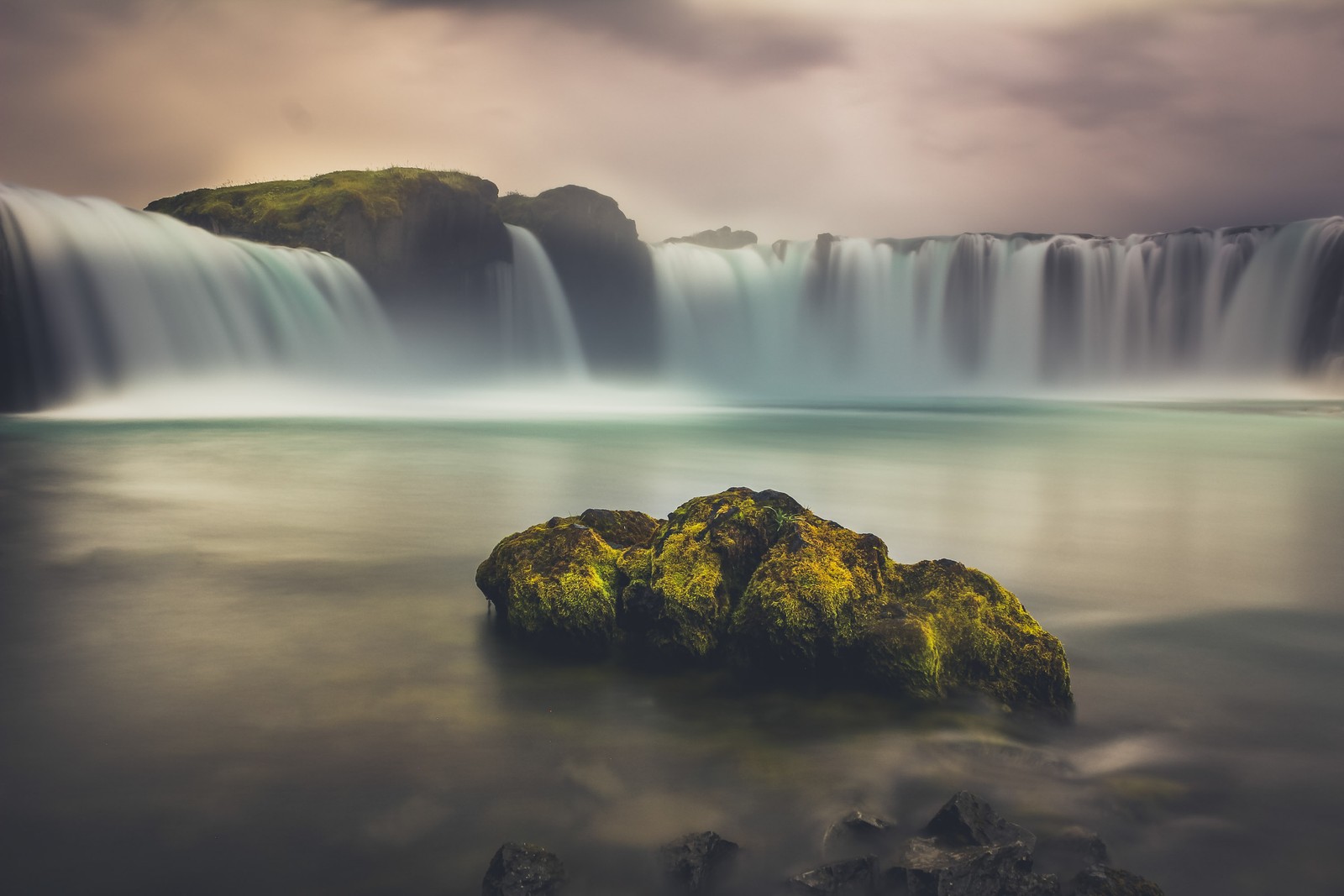 Uma grande cachoeira com uma rocha coberta de musgo em primeiro plano (goðafoss, cachoeira, islândia, cênico, paisagem)