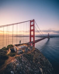 Ponte Golden Gate ao amanhecer, com vista para o oceano e uma figura solitária no penhasco.