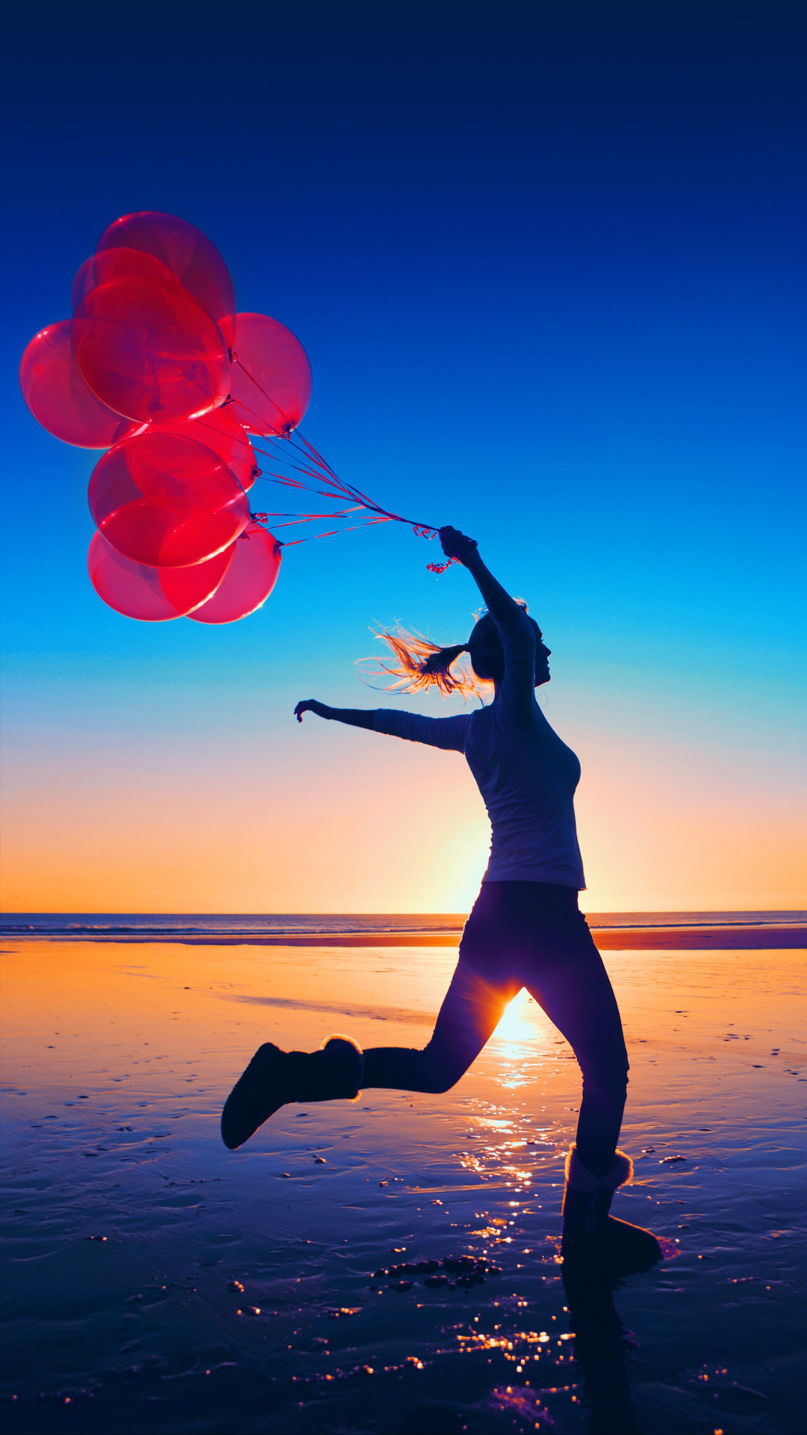 Une femme sautant avec des ballons au coucher du soleil sur la plage sous un ciel bleu (ballons, plage, running girl, été, coucher de soleil)