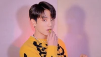 Jungkook do BTS em um suéter amarelo vibrante, segurando brincando uma uva com um olhar marcante.