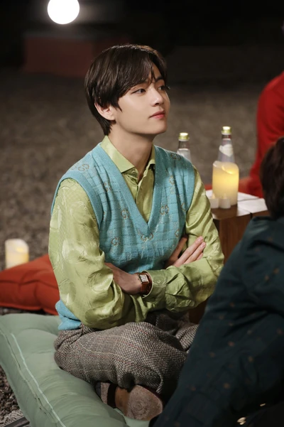 Taehyung in a sweater vest, seated with a thoughtful expression during a cozy gathering.