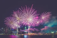 Fuegos artificiales vibrantes iluminan el cielo nocturno sobre el horizonte de la ciudad, celebrando una ocasión festiva con explosiones de luz rosa y púrpura.