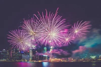 Fogos de artifício vibrantes iluminam o céu noturno sobre o horizonte da cidade, celebrando uma ocasião festiva com explosões de luz rosa e roxa.