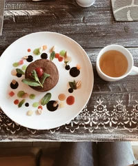 Plato de desayuno gourmet con un postre de chocolate adornado con menta y salsas coloridas, acompañado de una taza de té de hierbas en una mesa de madera rústica.