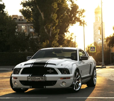 Ford Mustang blanche avec des rayures noires garée au premier plan, illuminée par la lumière du coucher de soleil, avec un clocher en arrière-plan.