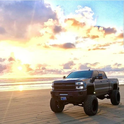 Camión Chevy negro estacionado en una playa de arena al atardecer, reflejando cielos vibrantes de color naranja y púrpura.