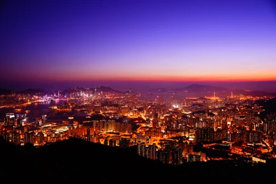 A breathtaking aerial view of Hong Kong's skyline at dusk, showcasing vibrant city lights against a gradient of deep blue and orange hues in the clear sky.