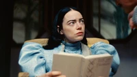 Emma Stone als Charakter in "Arme Dinge", nachdenklich blickend, während sie ein Buch hält, gekleidet in einem auffälligen blauen Outfit mit voluminösen Ärmeln.