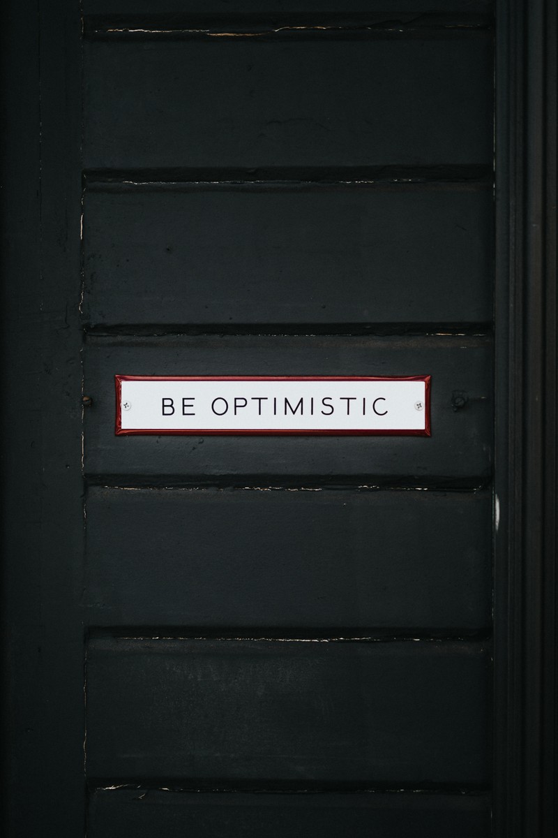 На двери есть знак, на котором написано «будь оптимистом» (be optimistic, вдохновляющие цитаты, тёмный фон, деревянная дверь, мотивационный)