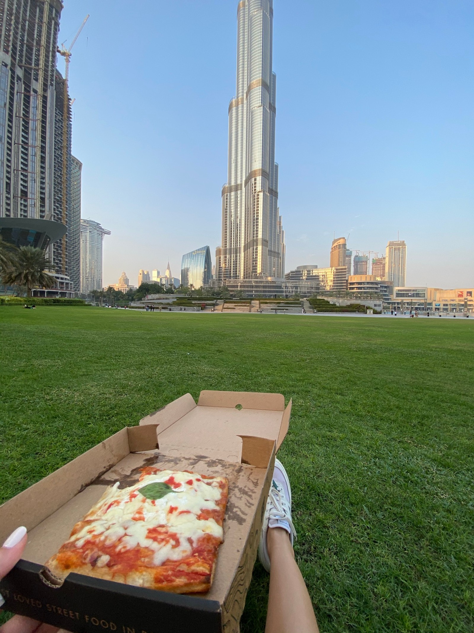 Someone is holding a box of pizza in front of a building (dubai, burj khalifa, urban area, skyscraper, daytime)