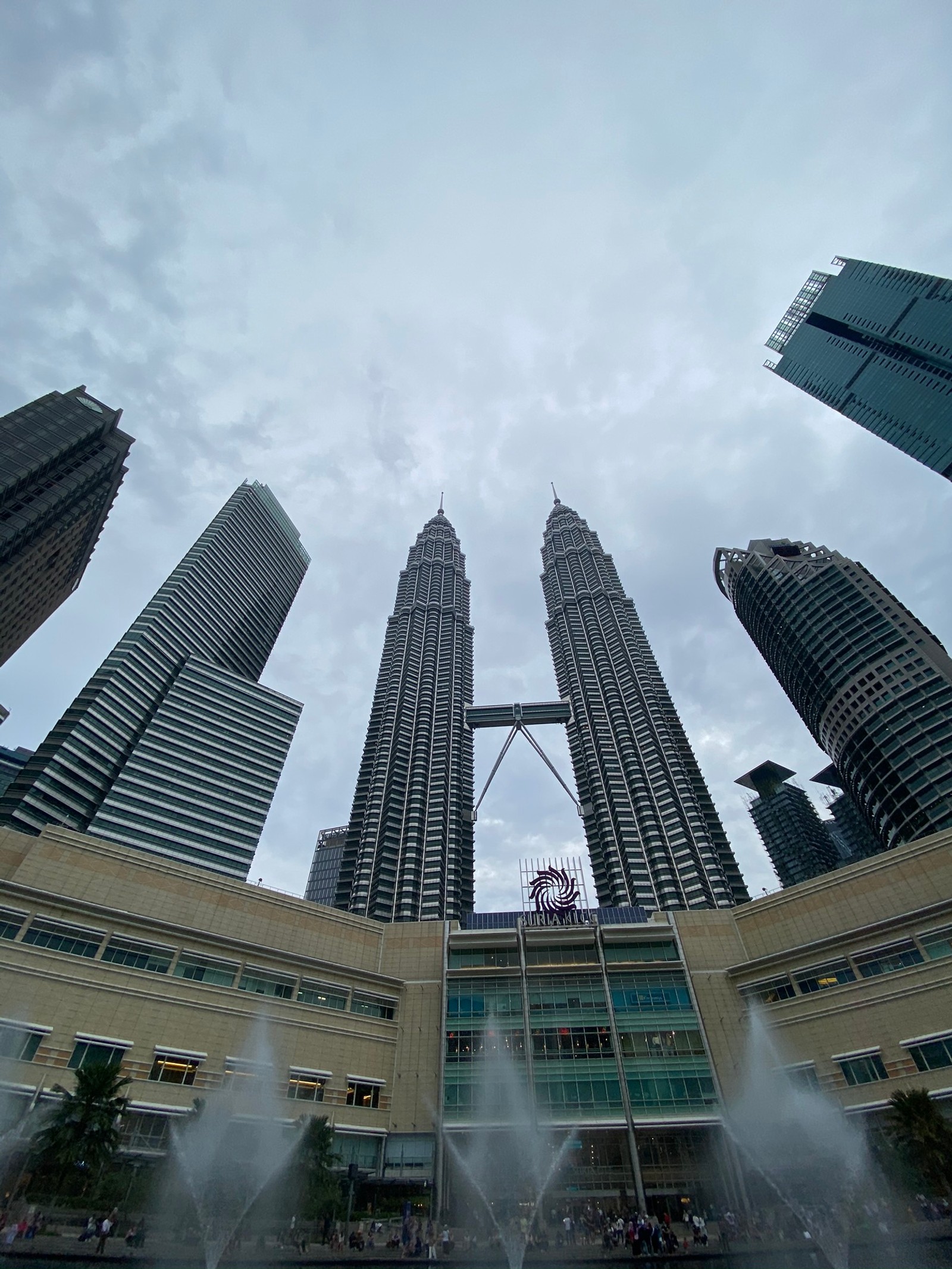 Há muitos edifícios altos ao fundo com uma fonte à frente (torres petronas, kuala lumpur, bloco de torre, dia, fonte)