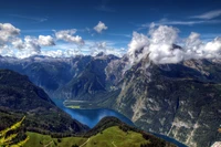 Deslumbrante vista aérea do Lago Königssee, situado nos Alpes da Baviera, cercado por montanhas majestosas e nuvens brancas, exibindo uma impressionante paisagem de verão.